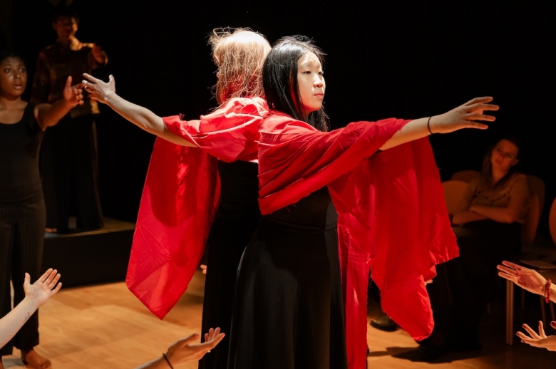 Two actors wearing bright red capes stand with their backs to each other with their arms outstretched in front of them.